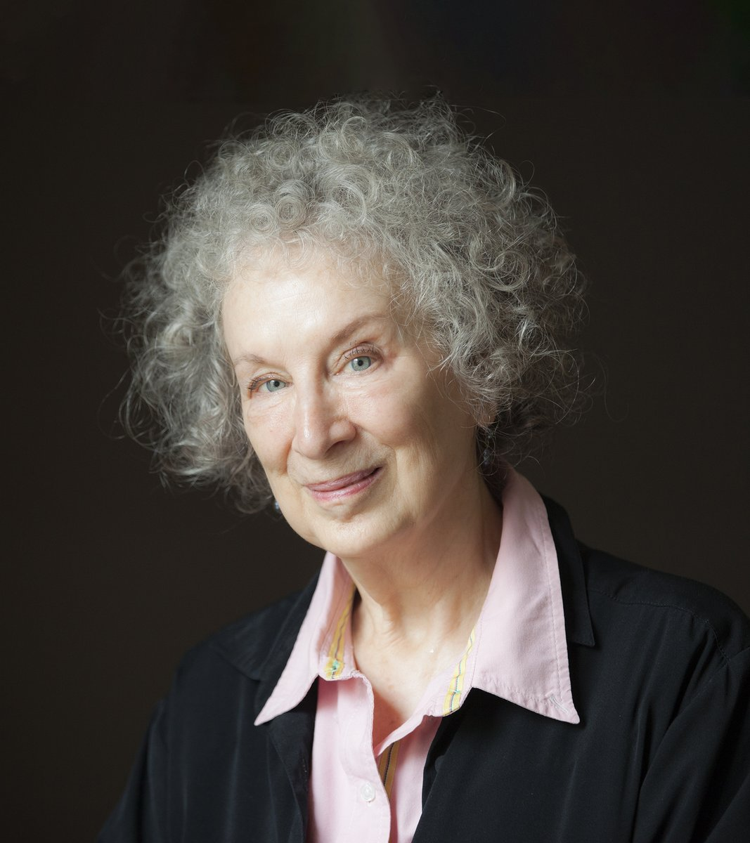 Una fotografía de Margaret Atwood. Se la ve alegre en la foto. Tiene el pelo rizado blanco y unos ojos azules impresionantes. Viste de camisa rosa y una chaqueta negra encima.